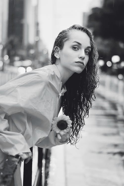 Free Black and white young woman with wet hair holding flower and looking on road in rainy day in city Stock Photo