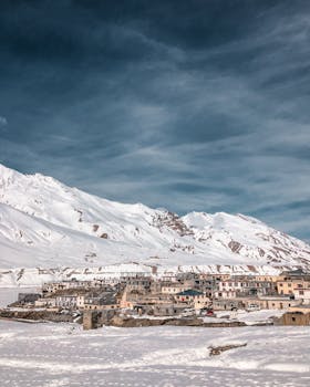 A stunning winter scene with snow-capped mountains and a quaint village beneath a cloudy sky.