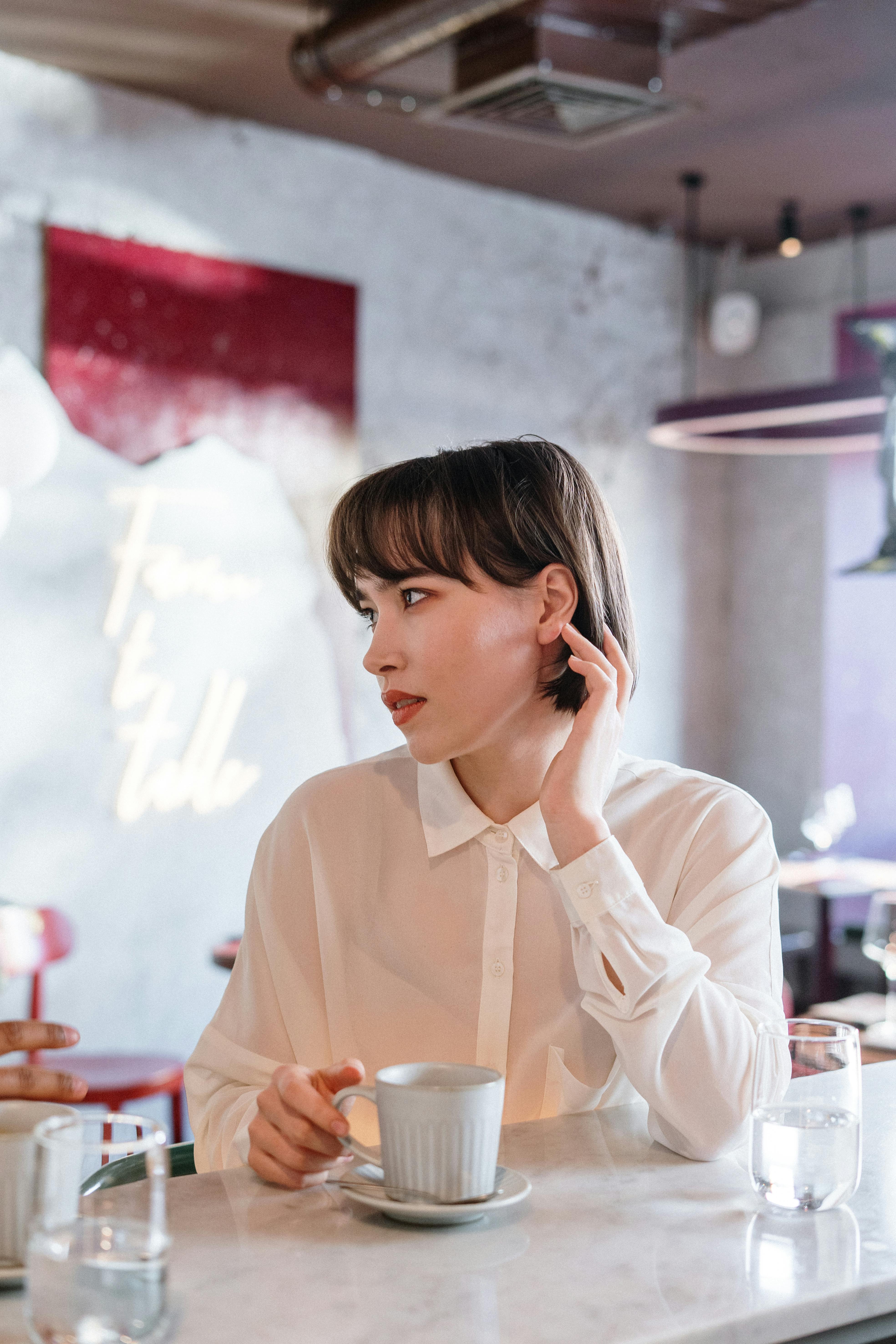 A stylish woman in a white blouse enjoys coffee at a modern indoor café setting.