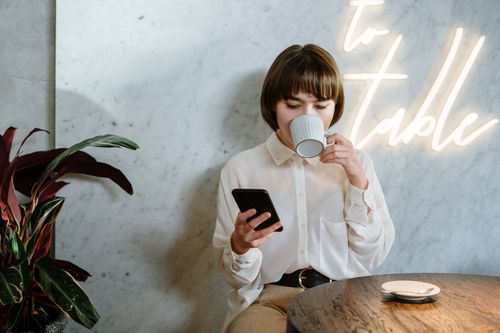 Free Stylish café scene with young woman enjoying her coffee, using a mobile phone under neon light ambiance. Stock Photo
