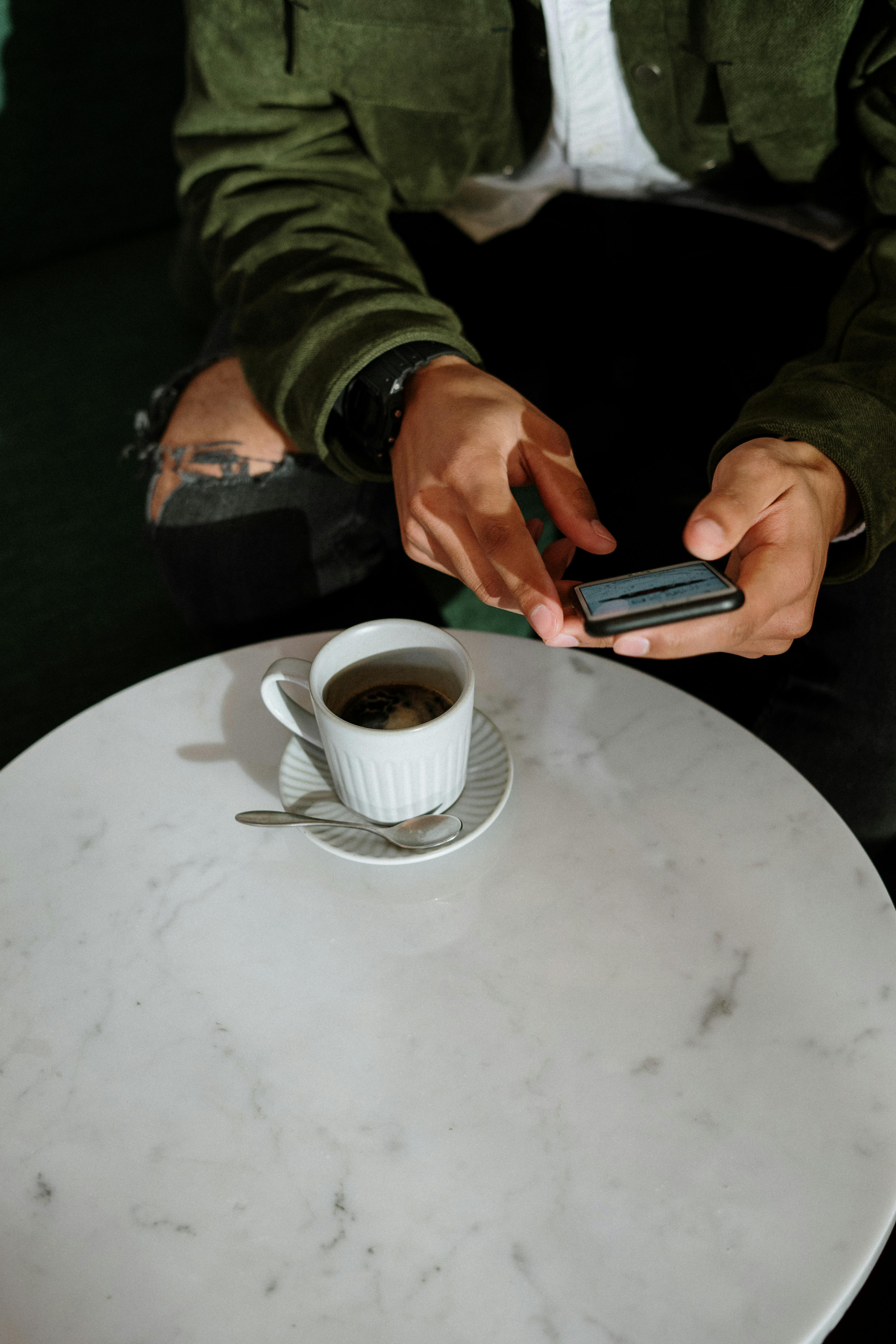 person holding black smartphone near white ceramic cup on white table