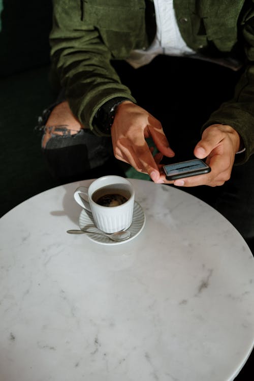 Person Holding Black Smartphone Near White Ceramic Cup on White Table