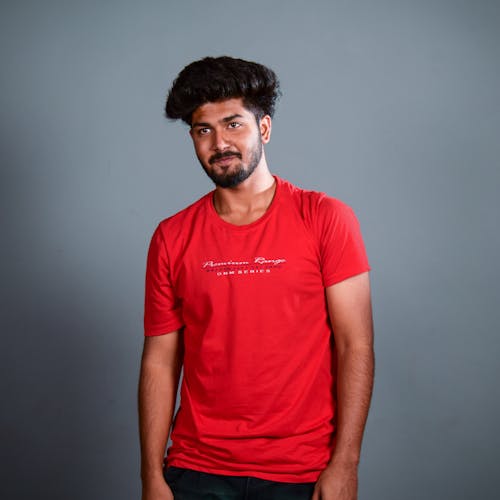 Young ethnic man with beard and bushy hair wearing red t shirt and looking away on gray background