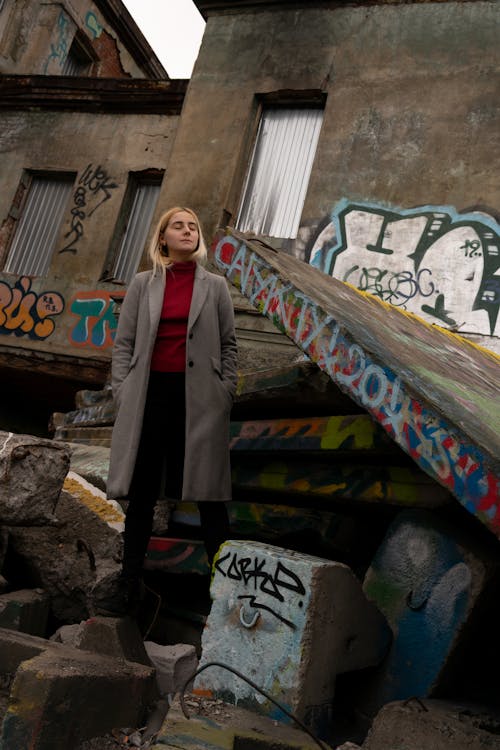 Low angle of blond woman in gray coat standing on concrete blocks with graffiti around in gloomy day