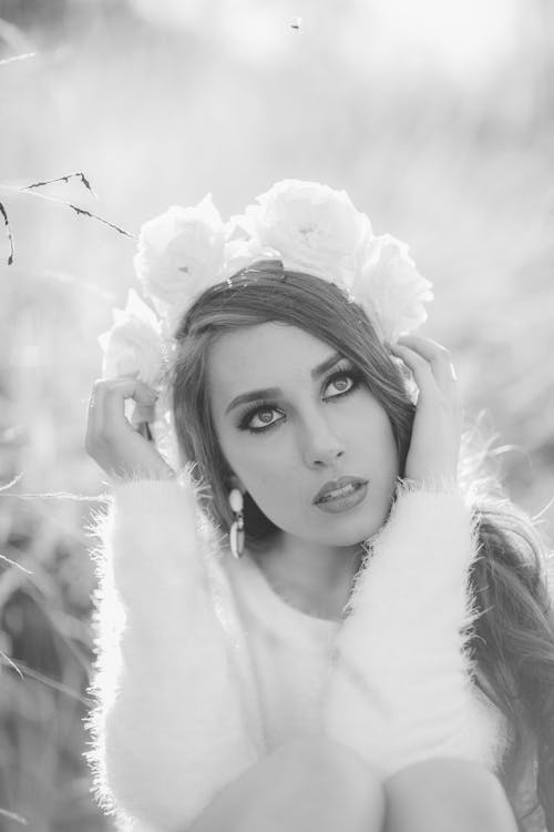 Free From above black and white of young dreamy woman with makeup in earring and fluffy sweater looking up while touching decorative wreath on head Stock Photo