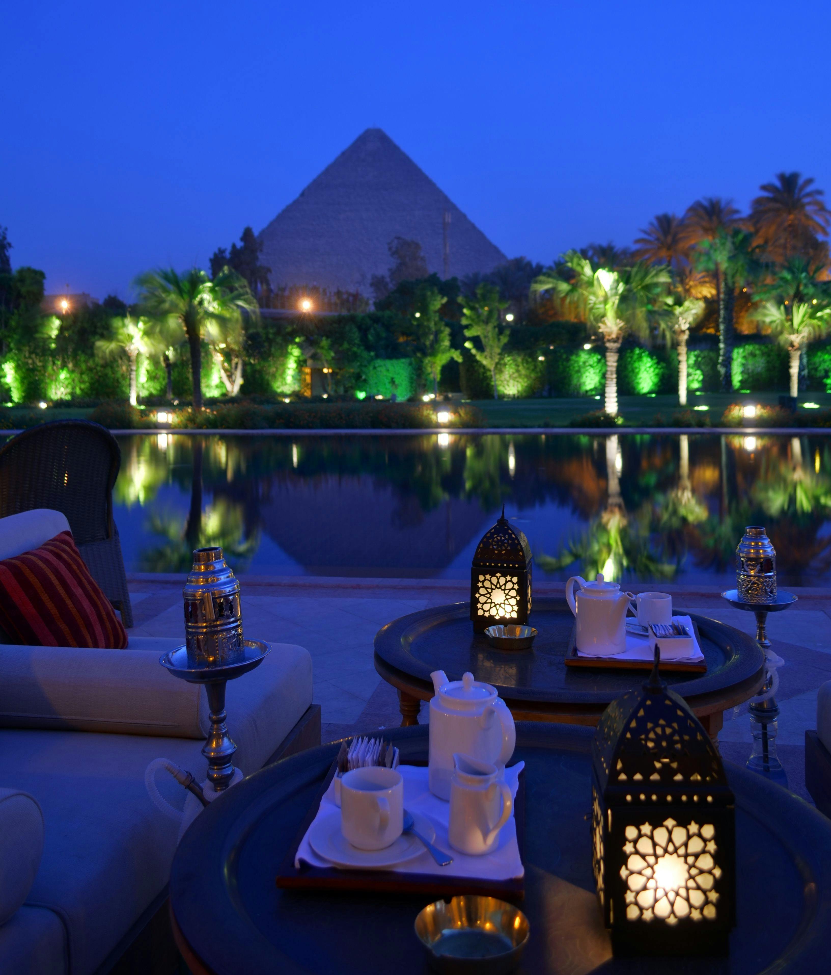 From above of cafe with dishware and ornamental lanterns on round tables near sofa and pond reflecting trees and old pyramid under blue sky illuminated by artificial lights