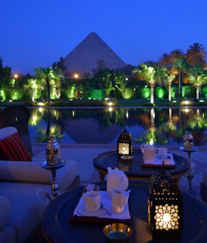 From above of cafe with dishware and ornamental lanterns on round tables near sofa and pond reflecting trees and old pyramid under blue sky illuminated by artificial lights