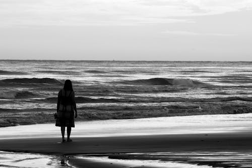 Black and white back view of unrecognizable alone female silhouette admiring stormy ocean while standing on sandy coast under cloudy sky with horizon line