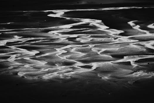 From above black and white scenery view of sandy ocean coast with wavy water shining on surface at night