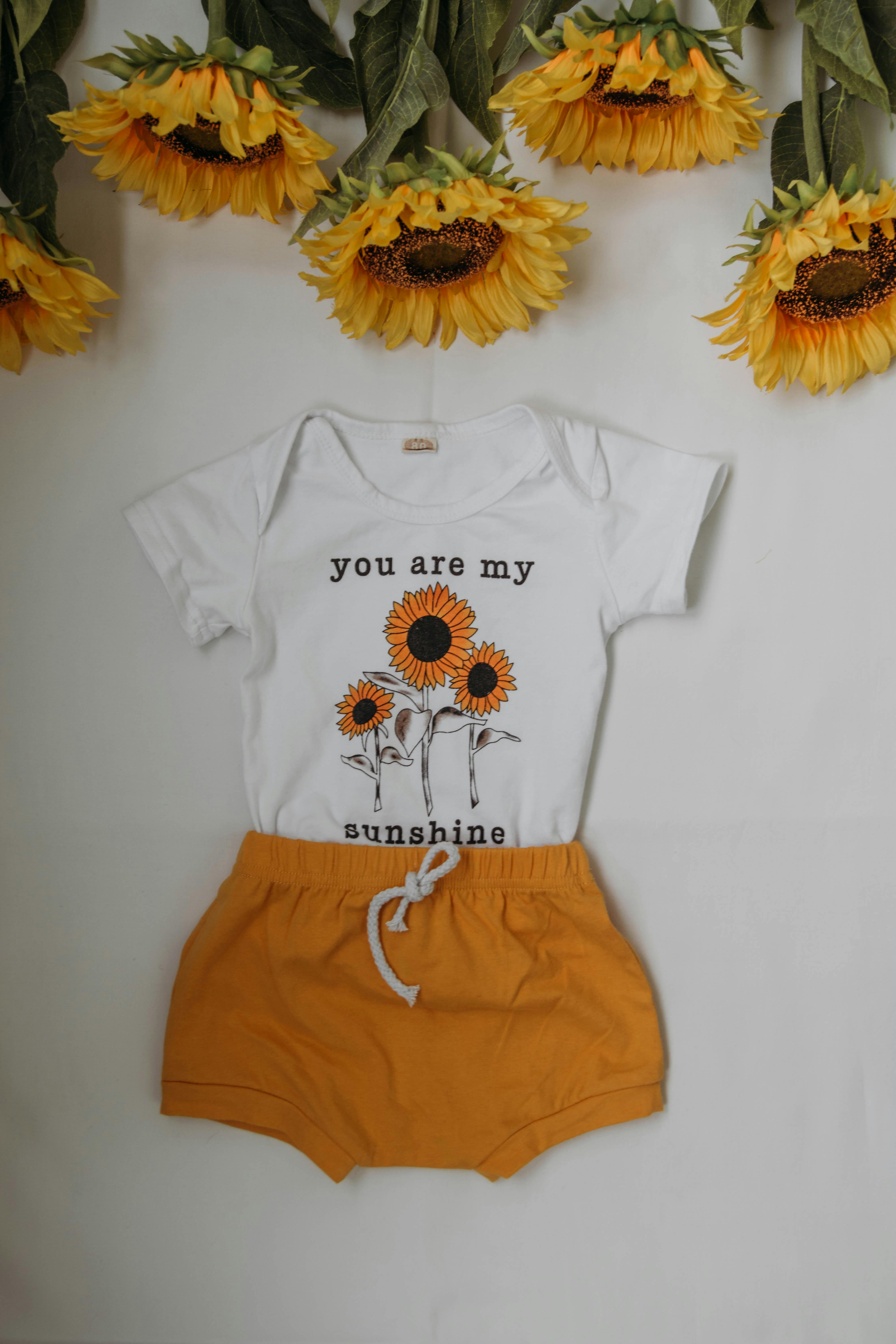 From above of kid home wear with orange shorts and white t shirt near colorful sunflowers on white table