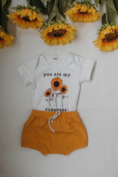 From above of kid home wear with orange shorts and white t shirt near colorful sunflowers on white table