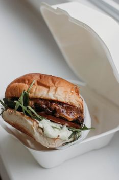 From above of tasty burger with soft buns and juicy meat with barbecue sauce above fresh rosemary leaves and melted cheese in plastic box on windowsill in flat