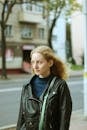 Young emotionless blond female in leather jacket looking away while standing on sidewalk near asphalt roadway and house facade in town