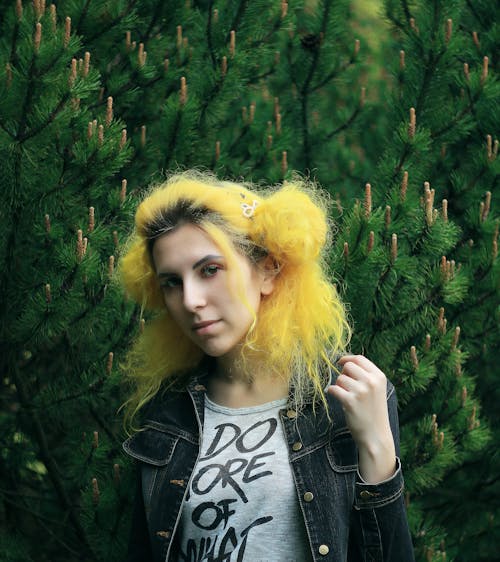 Young stylish ethnic female in casual wear with dyed yellow hair standing near pine tree while looking at camera