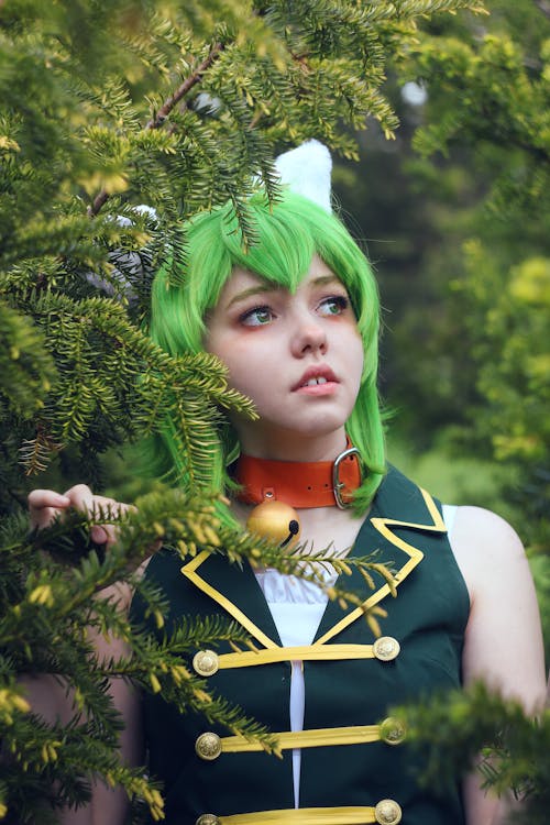 Pondering female teenager in costume and colorful wig with evil horns looking away while standing near coniferous tree in daytime