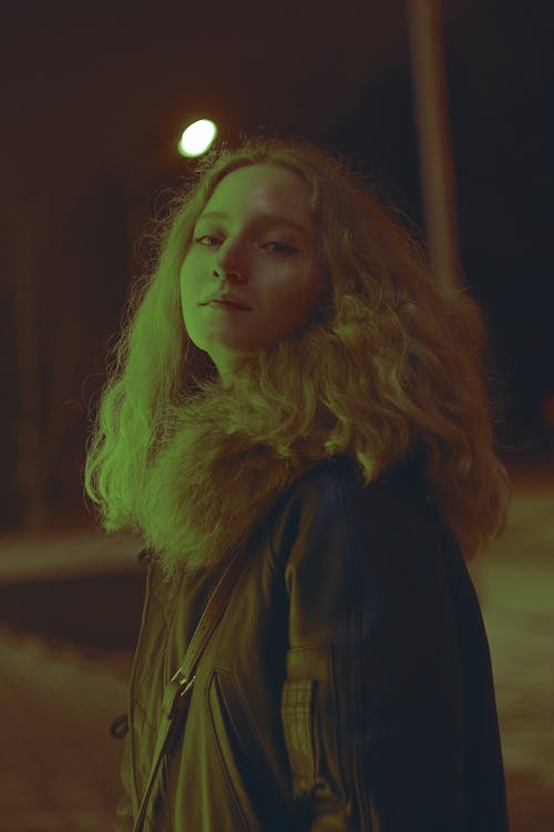 Side view of trendy blond female in outerwear with fur collar looking at camera while standing on street near glowing lamp post at night