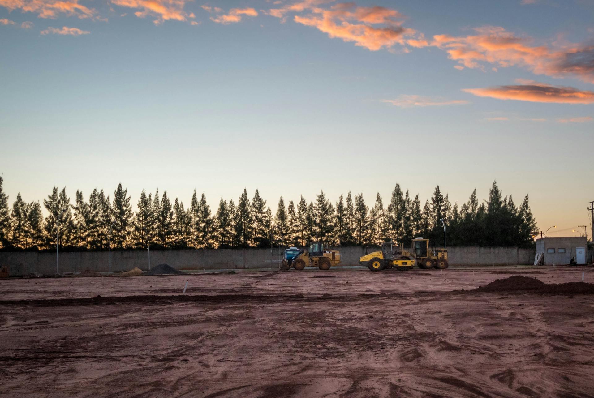 Heavy machinery on fenced site construction with forest under sky with clouds in evening time