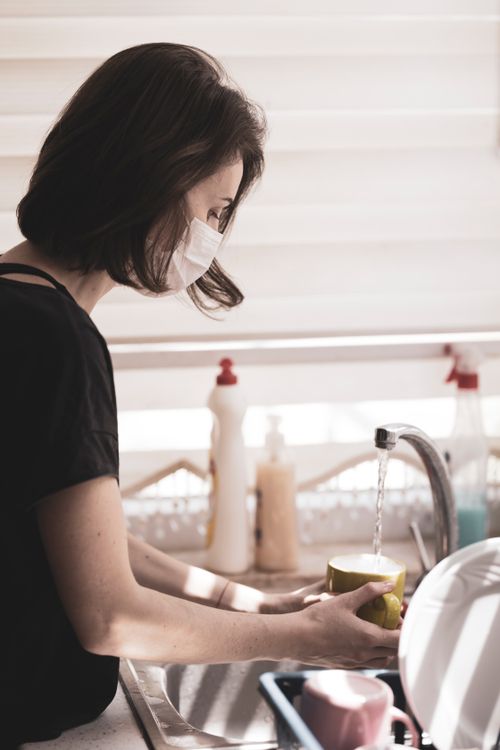 Free Woman with face mask washing dishes at a kitchen sink, reflecting new normal hygiene practices. Stock Photo