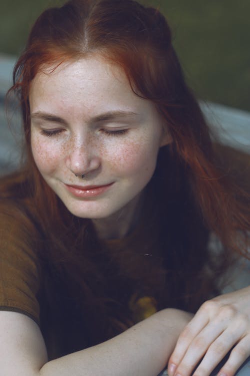 Positive young female with freckles and red hair smiling and sitting with closed eyes