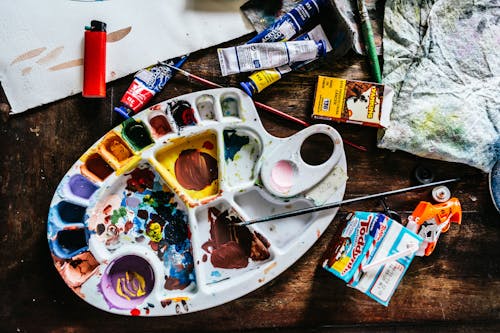 Top view of various art supplies including paintbrushes with colorful tubes of paint and palette placed on wooden table