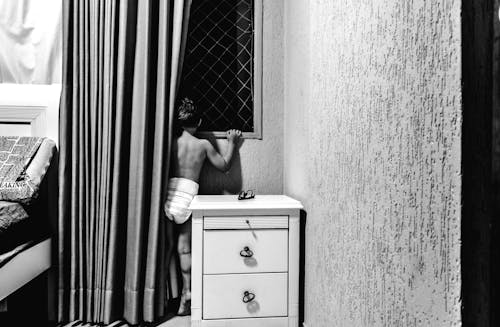 Black and white back view of anonymous boy in underwear standing in bedroom near curtains and looking through window