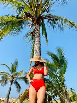 From below of female in stylish red swimwear and hat standing near palm tree under blue sky in summer day and looking away