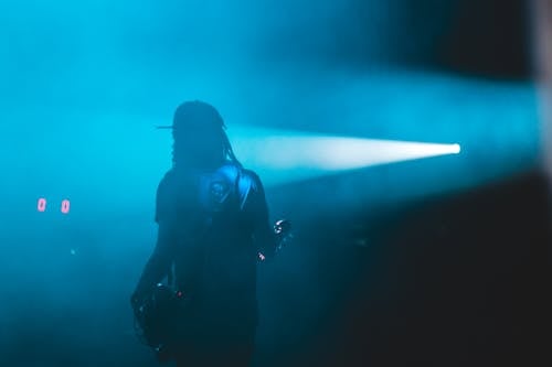 Back view of anonymous male in cap standing near bright spotlight in dark place