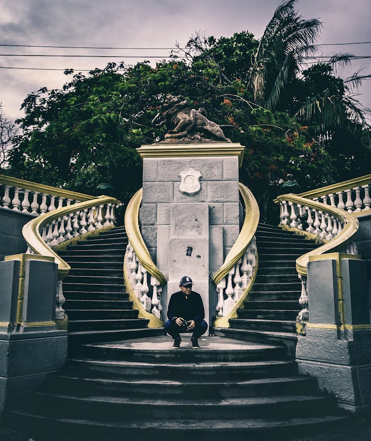 Man Sitting On Squat On Stairs Near Monument In Park