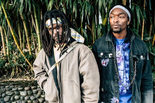 Calm confident African American males with dreadlocks wearing street style clothes and headbands standing against tall green trees in garden