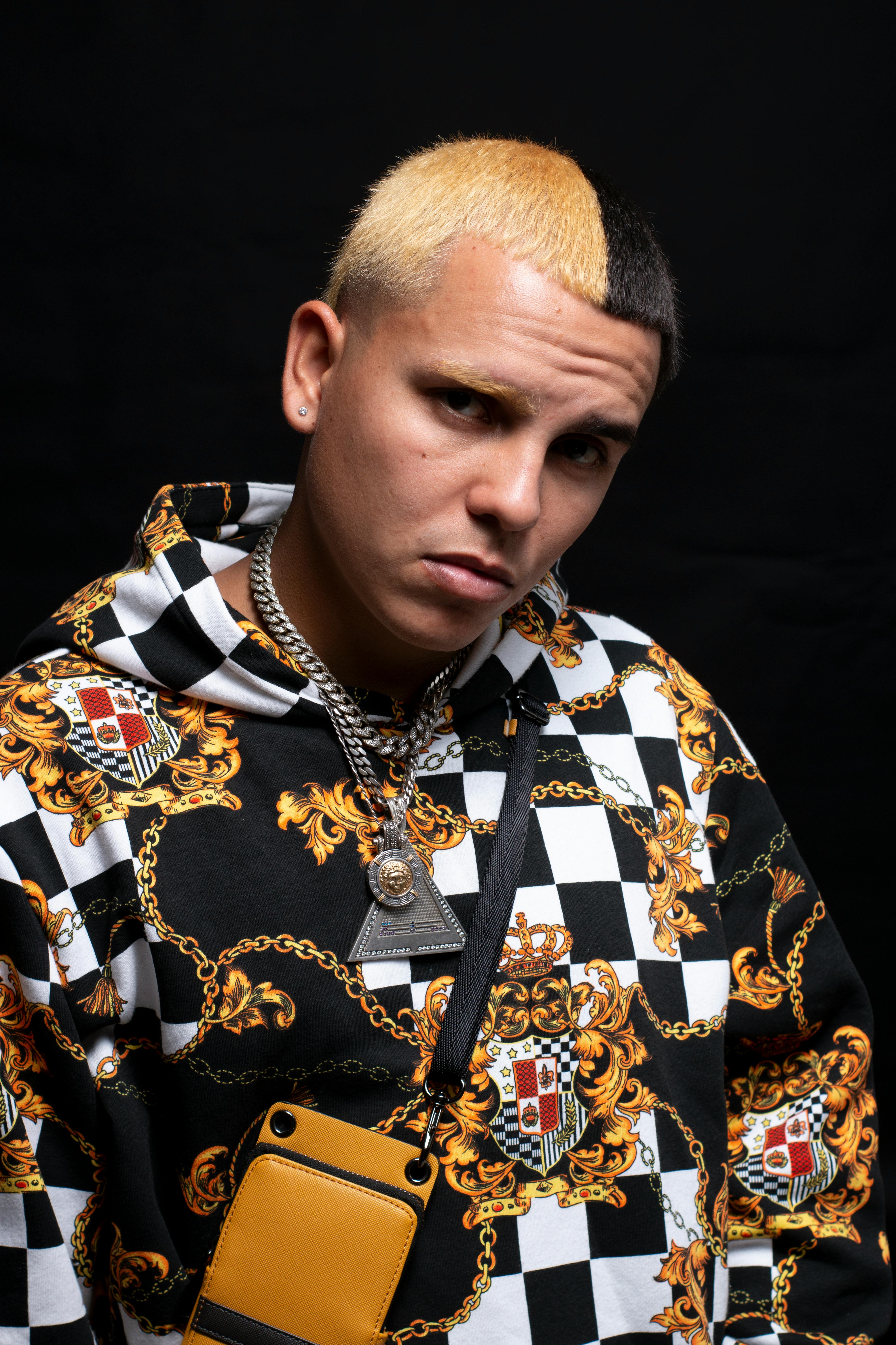 Stylish young man in vibrant patterned hoodie and jewelry posing with confidence in a studio setting.