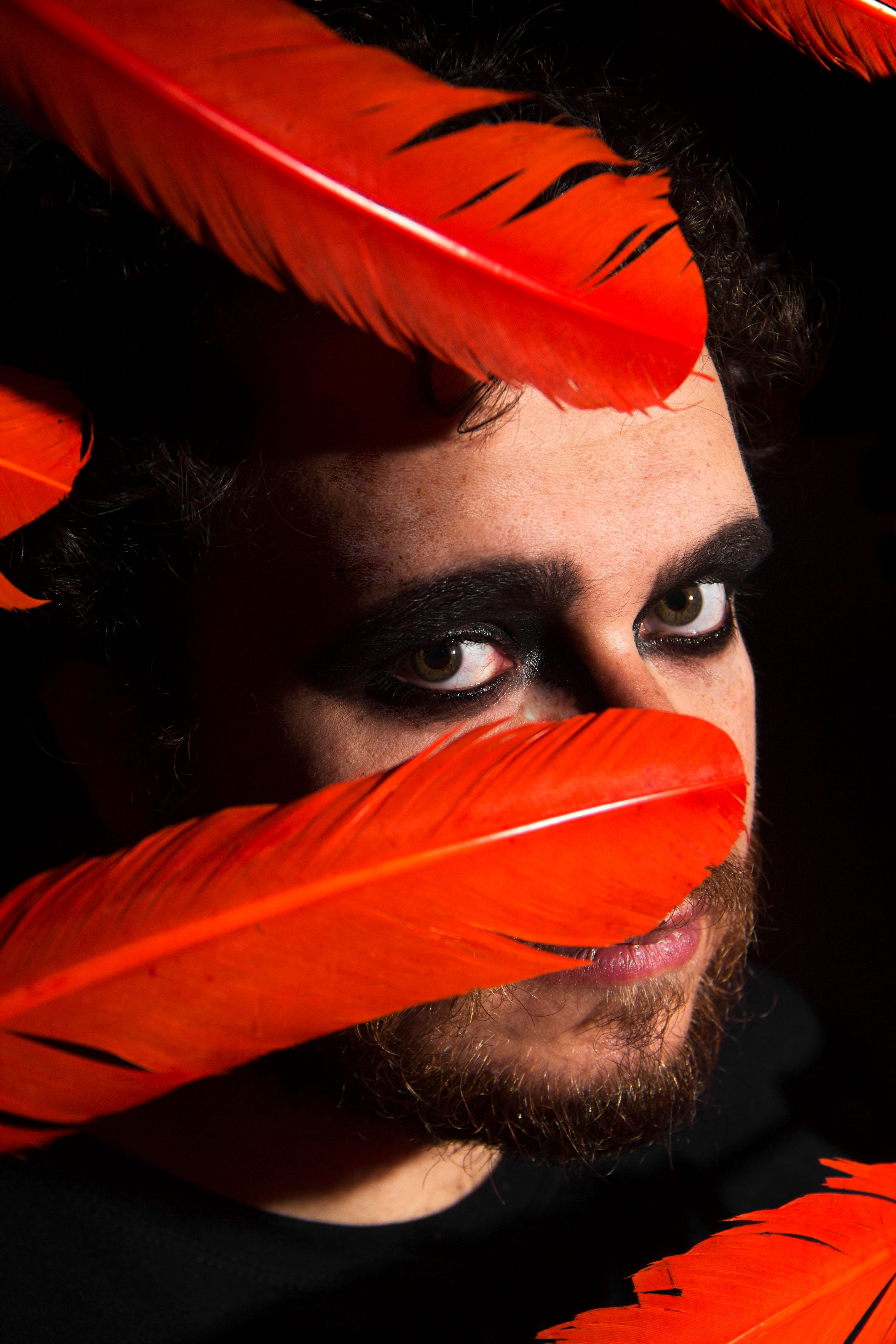 Creative studio shot of calm bearded male with eyes makeup behind red feathers on black background