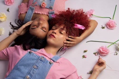 Free From above young serene ethnic females in similar casual wear lying on floor with eyes closed amidst delicate flowers and touching each others heads Stock Photo
