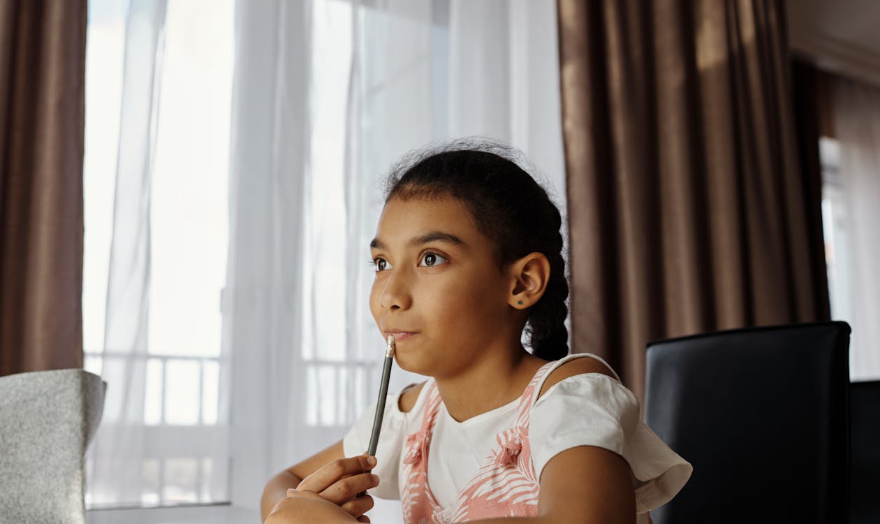 Young girl holding pencil, looking pensive while doing homework at desk