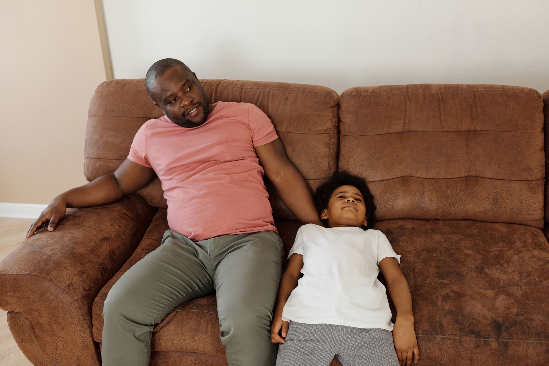 A father and son lounging comfortably on a brown sofa in a cozy indoor setting.