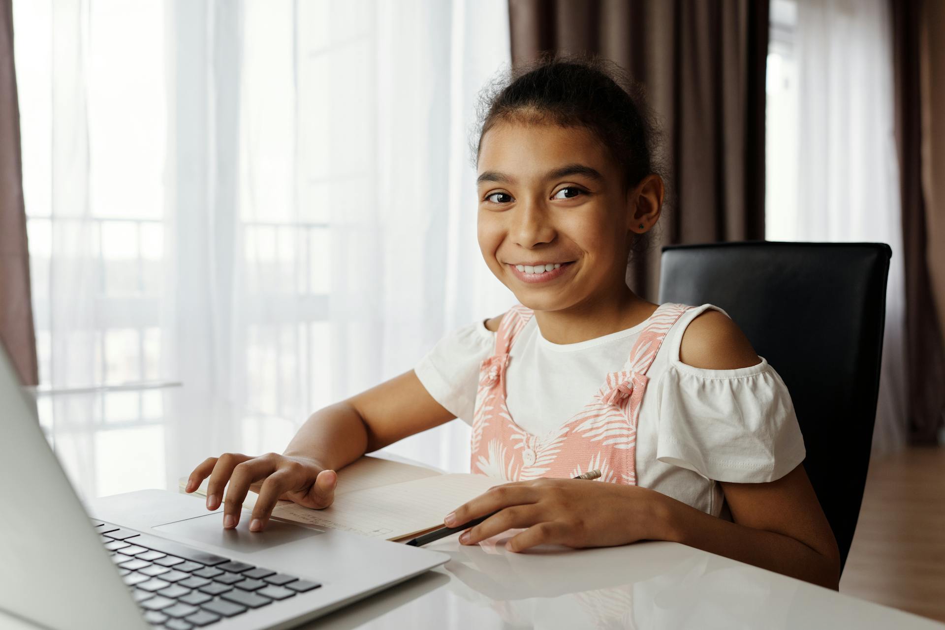 A cheerful young girl sits at a desk using a laptop for online learning at home.