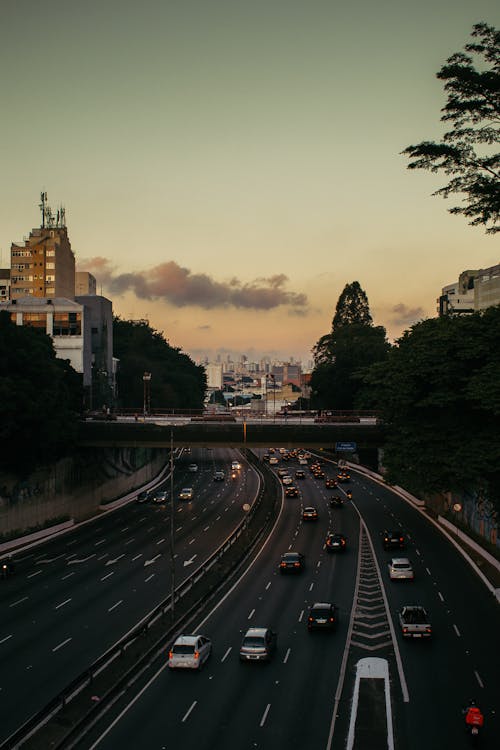 Various vehicles riding along asphalt road near pedestrian bridge and buildings in modern city during sunset