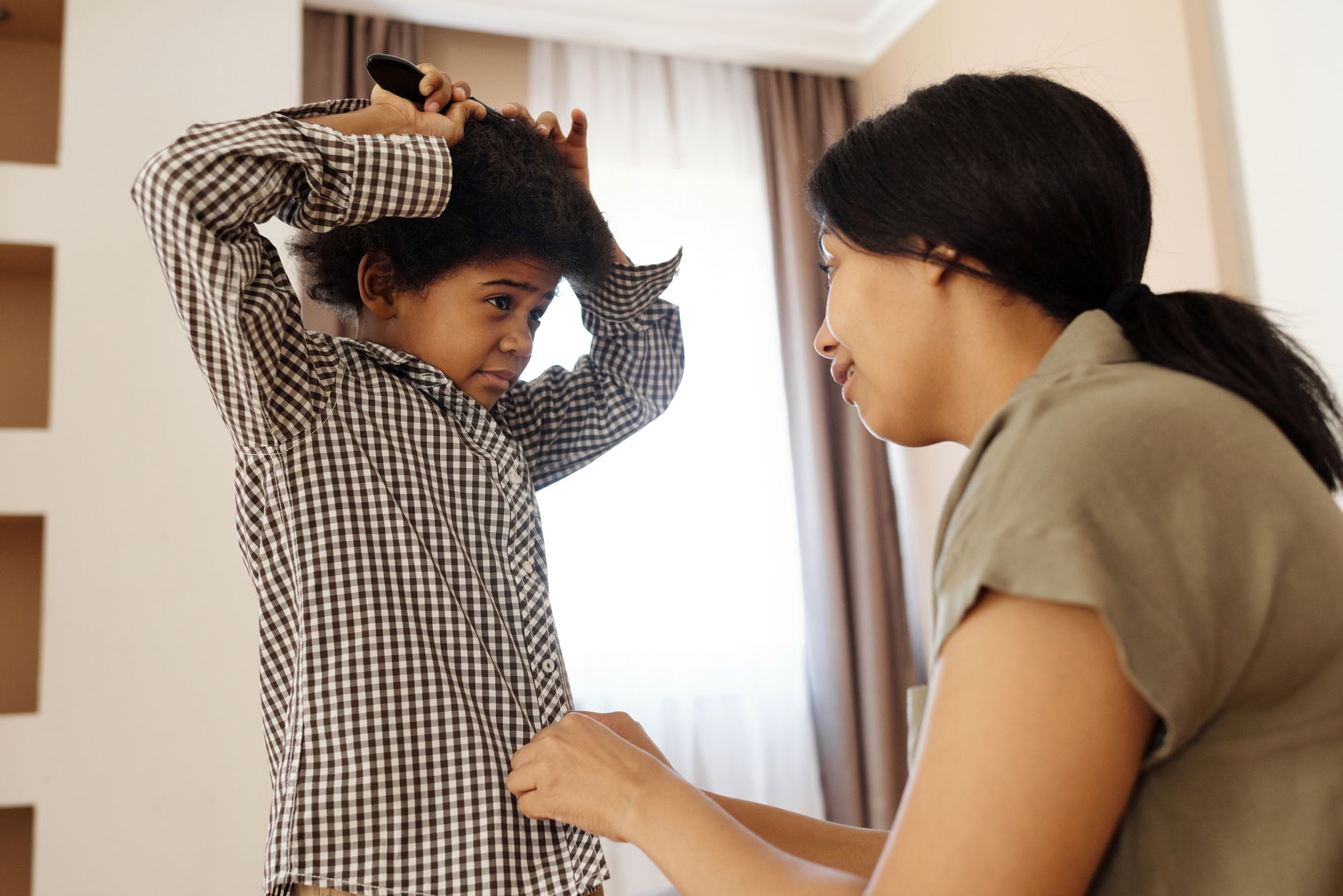A caring mother helps her son brush his hair and button his shirt indoors.