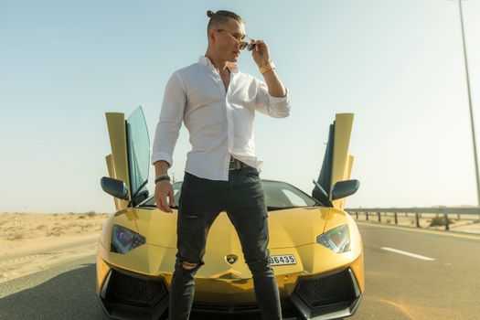 Young confident male with stylish haircut in white shirt and jeans standing near yellow sports car and adjusting sunglasses