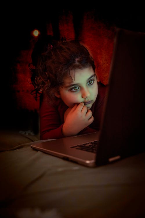 Concentrated girl in casual clothing with dark hair tied up browsing internet on laptop while resting in dark room