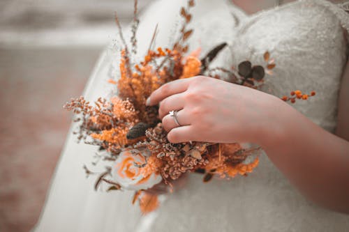 Crop fiancee in white wedding dress with ring on finger touching yellow delicate flowers in wedding bouquet
