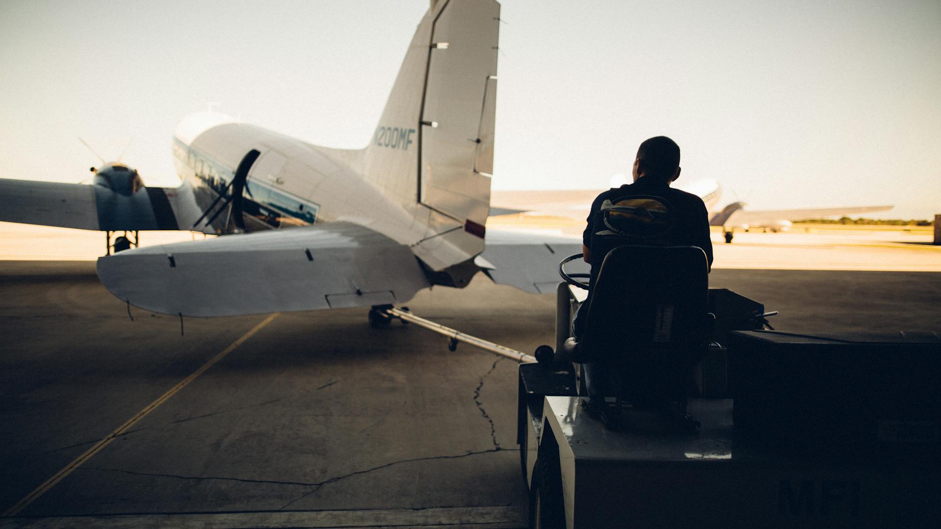 Silhouette of worker driving baggage carrier on paved airfield with airplane before flight