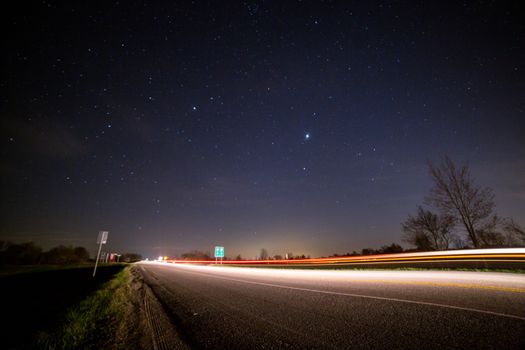 From below of glowing asphalt road with fence near trees and road signs under picturesque night sky with luminous stars at sundown