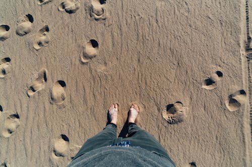 Free From above of crop unrecognizable barefoot male in jeans standing on sandy shore with loose texture and deep holes in summer Stock Photo