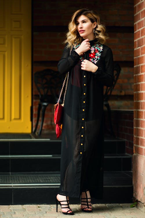 Woman in Black Long Sleeve Dress Standing Beside Brick Wall