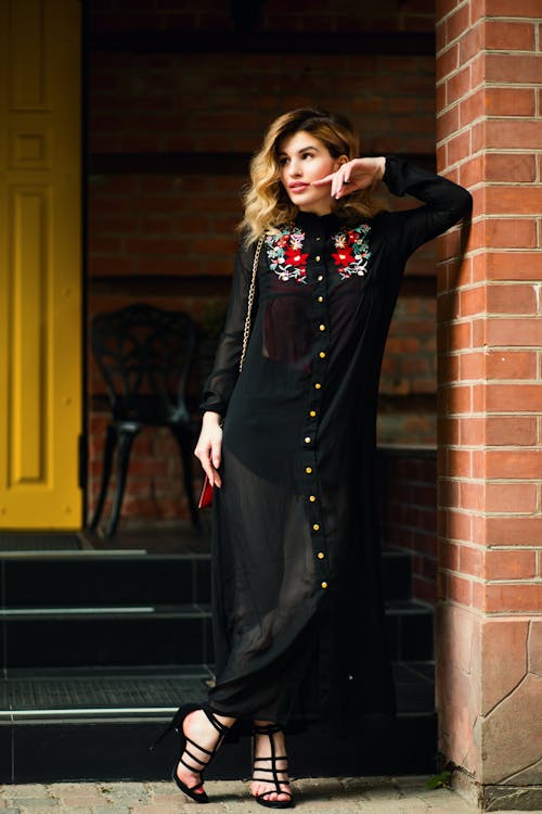 Full body of trendy female wearing long black outfit leaning on brick wall and looking away