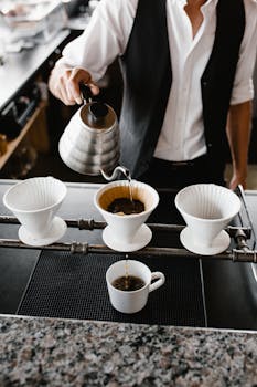 Barista pouring hot coffee using a kettle into a cup in a modern indoor coffee shop.