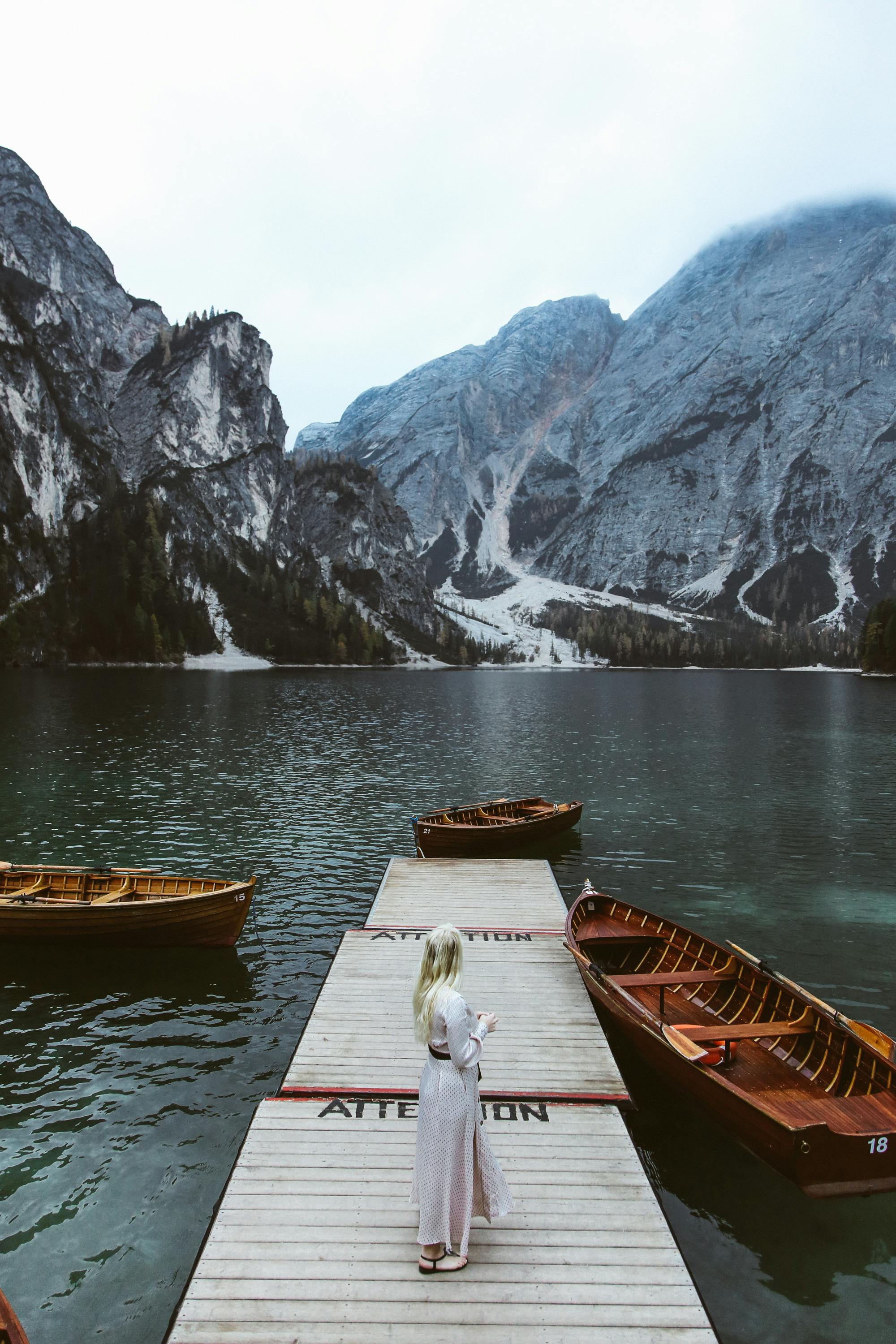 A woman stands on a dock surrounded by boats and stunning mountain and lake views.