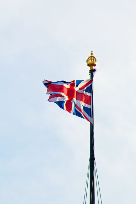 From below of wavy British flag with red cross on pointed stick with crown on top under cloudy sky
