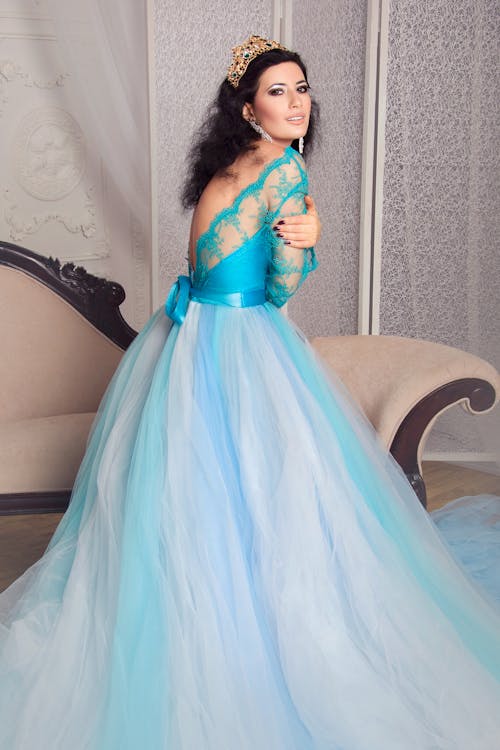 Side view elegant female in fancy maxi gown of blue color standing in classic interior while hugging self and looking at camera