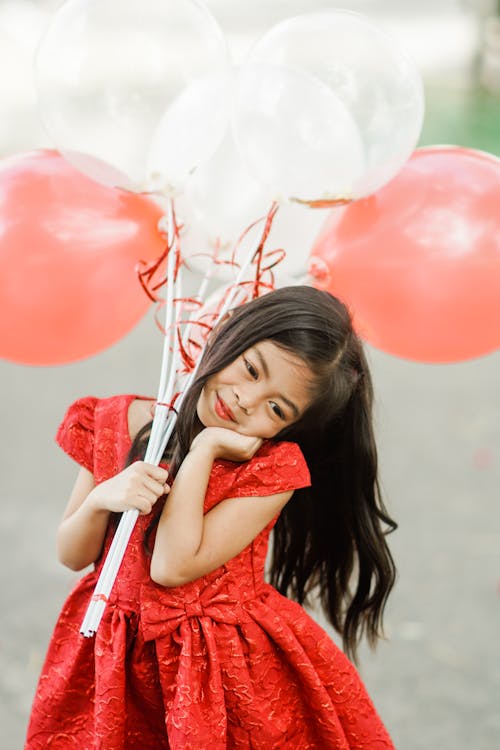 Cute Girl in a Red Dress Holding Red and White Balloons